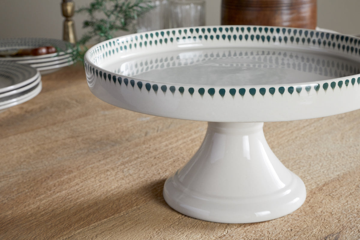 The nkuku Juniper Drop Ceramic Stand - Cream & Juniper features a green drop border and sits on a wooden table with stacked plates, glasses, and blurred decor in the background.