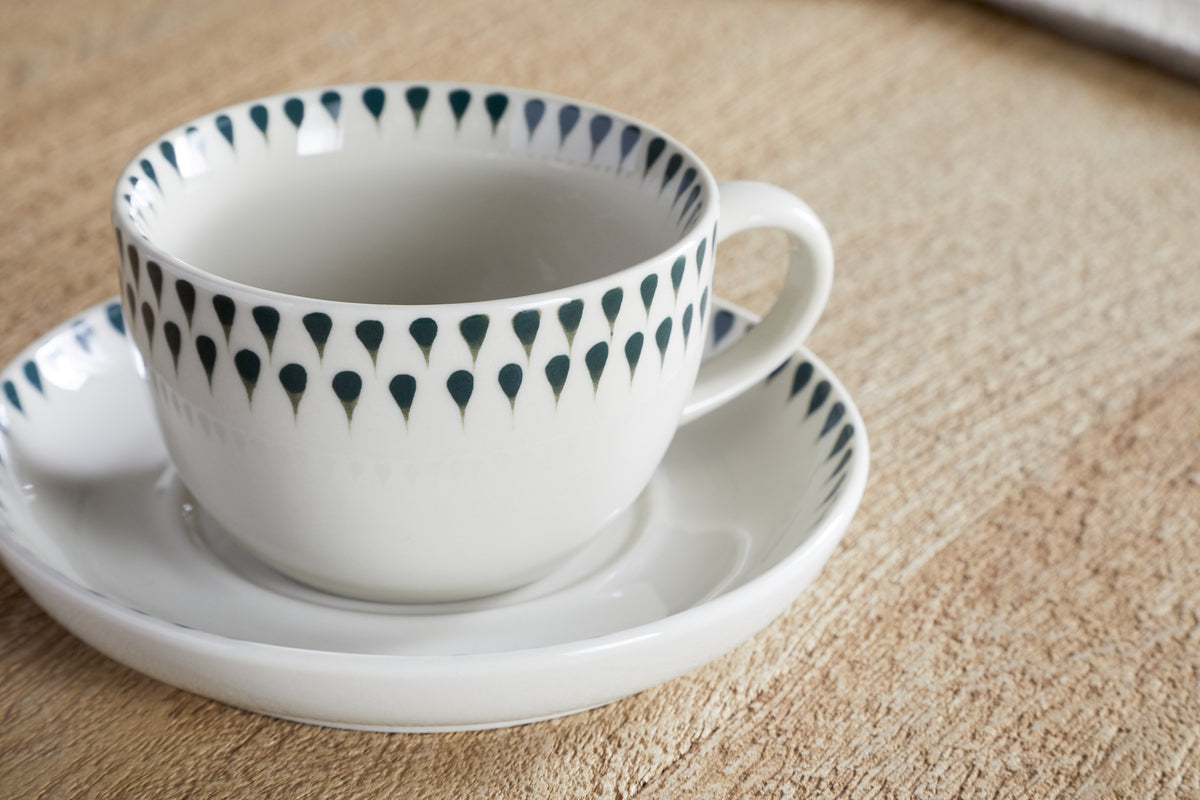 A nkuku Juniper Drop Cup & Saucer - Cream & Juniper, featuring a blue-green drop rim pattern, rests on its matching saucer atop a textured wooden surface.