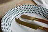 The nkuku Juniper Drop Dinnerware Set - Cream & Juniper (Set of 12) displays blue teardrop patterns on the rim and is shown holding two brass cheese knives with wooden handles, arranged on a textured surface with a white cloth backdrop.
