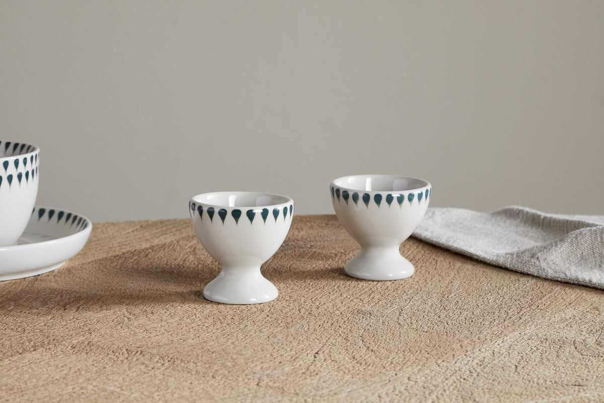 The Juniper Drop Egg Cups - Cream & Juniper by nkuku (set of 2) with blue teardrop patterns sit on a textured brown table next to a folded beige cloth. Matching plate and bowl partially visible on the left.