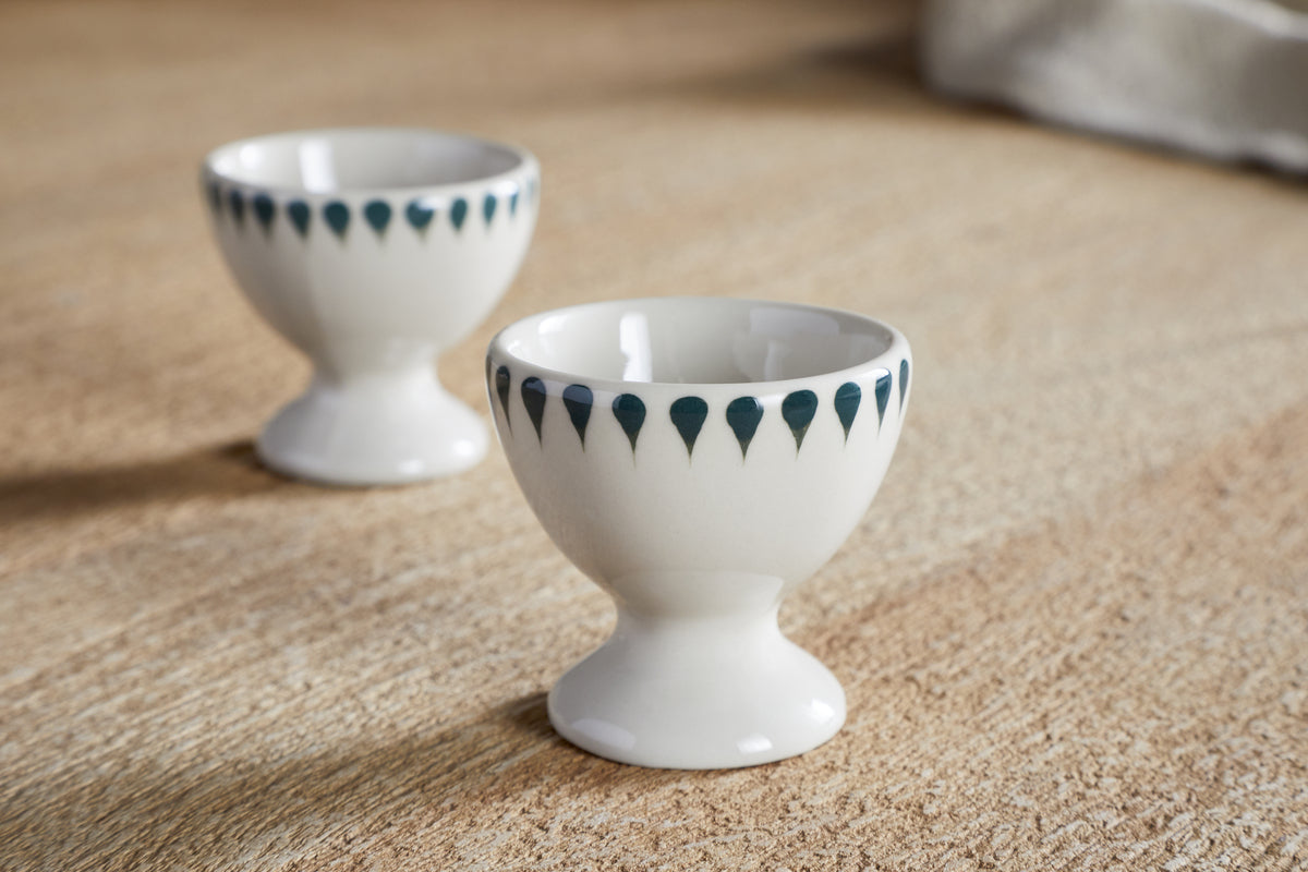 The nkuku Juniper Drop Egg Cups - Cream & Juniper (Set of 2) feature a dark blue teardrop pattern on white ceramic, ideal for adding style to your breakfast table.
