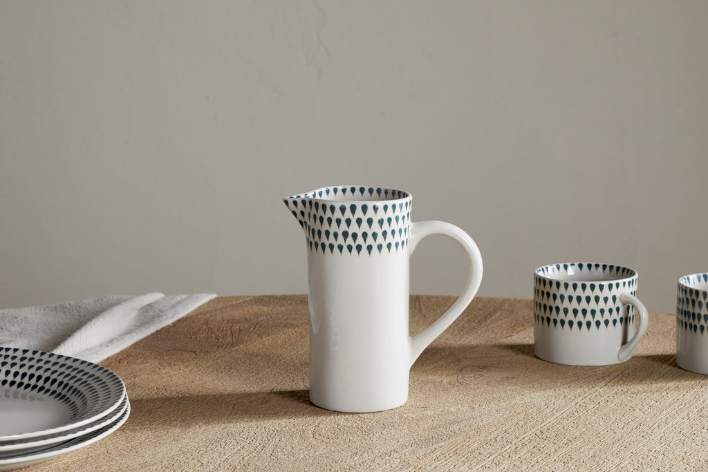 The nkuku Juniper Drop Jug - Cream & Juniper - Small sits on a wooden table, accompanied by matching cups and plates in the same dark teardrop pattern. A folded napkin is placed beside the plates on the left.