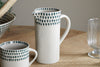 The nkuku Juniper Drop Jug - Cream & Juniper - Small features a handle and a teal teardrop rim, displayed on a wooden table beside its matching mug and a clear glass goblet—ideal for modern tableware settings.