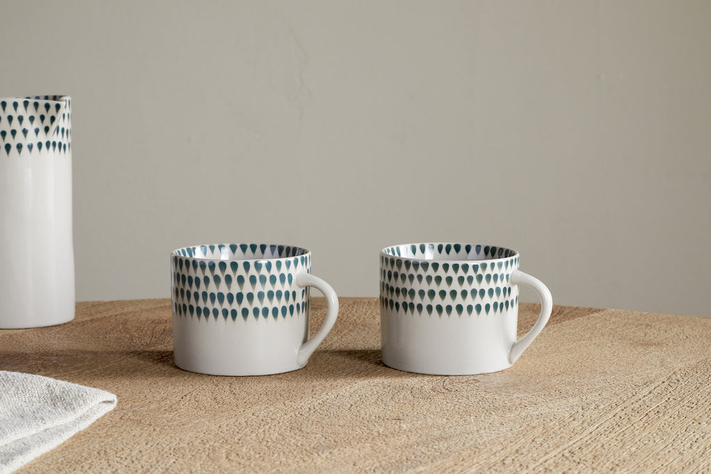 Two nkuku Juniper Drop Mugs in cream and juniper, featuring unique handles and signature drop design, rest side by side on a wooden surface. A matching vase and a white cloth are partially visible to the left. Set of 2 (Small).
