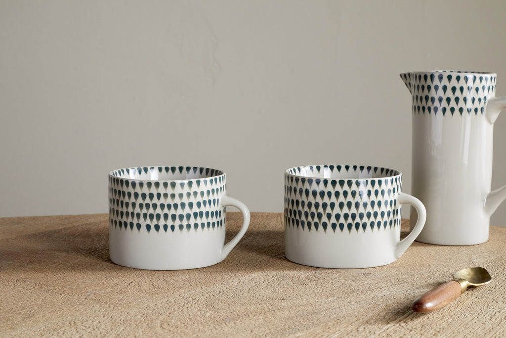 Two nkuku Juniper Drop Mugs – Cream & Juniper, Large (Set of 2) rest on a wooden surface beside a matching pitcher and a wooden-handled spoon, with a simple neutral background.