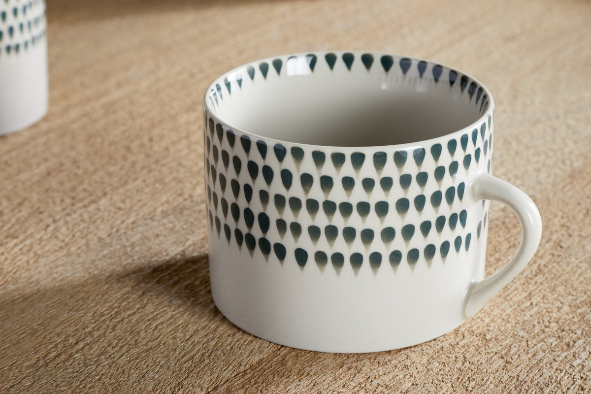 The nkuku Juniper Drop Mug - Cream & Juniper - Large (Set of 2) features a white ceramic body, dark blue teardrop pattern near the rim, simple handle, and glossy finish, shown on a wooden surface.