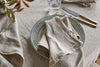 A table set with a beige linen tablecloth, patterned plate, Junto Linen Napkin - Natural by nkuku, gold-handled cutlery, and a clear glass. Red-green plant leaves are visible in the corner.