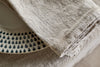 A close-up of the nkuku Junto Linen Napkin - Natural (Set of 4) draped over a table edge, partially covering a white plate with dark blue teardrop patterns.