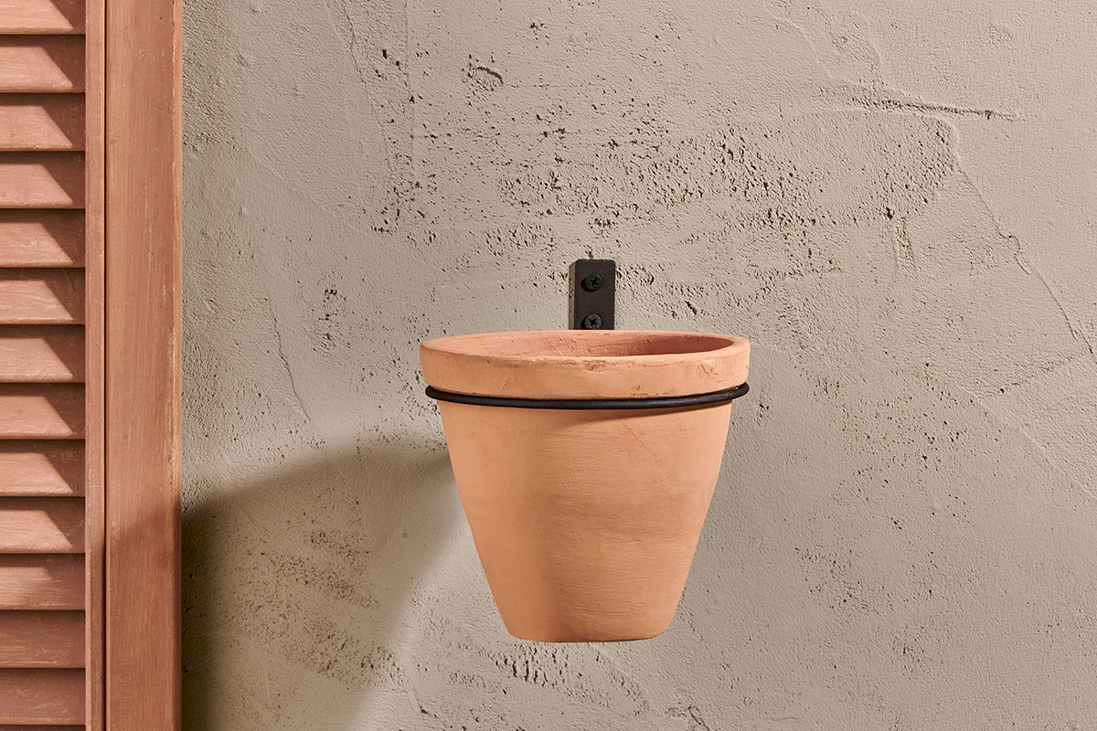 The nkuku Jara Single Wall Hung Planter, a terracotta pot with a black metal bracket, is mounted on a beige textured wall. A wooden shutter appears to the left, and the empty planter casts a gentle shadow on the serene wall.