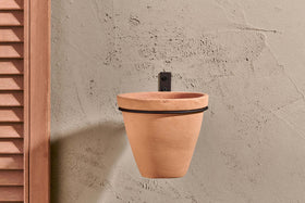 The Jara Single Wall Hung Planter by nkuku, a plain terracotta pot in a black metal holder, hangs on a textured beige wall. To the left is part of a wooden shutter, and the empty planter casts a faint shadow on the wall.