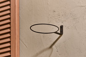 The nkuku Jara Single Wall Hung Planter, a round metal ring, is mounted horizontally on a beige textured wall with a bracket and two screws beside a wooden louvered panel. Soft natural light casts subtle shadows over the display.