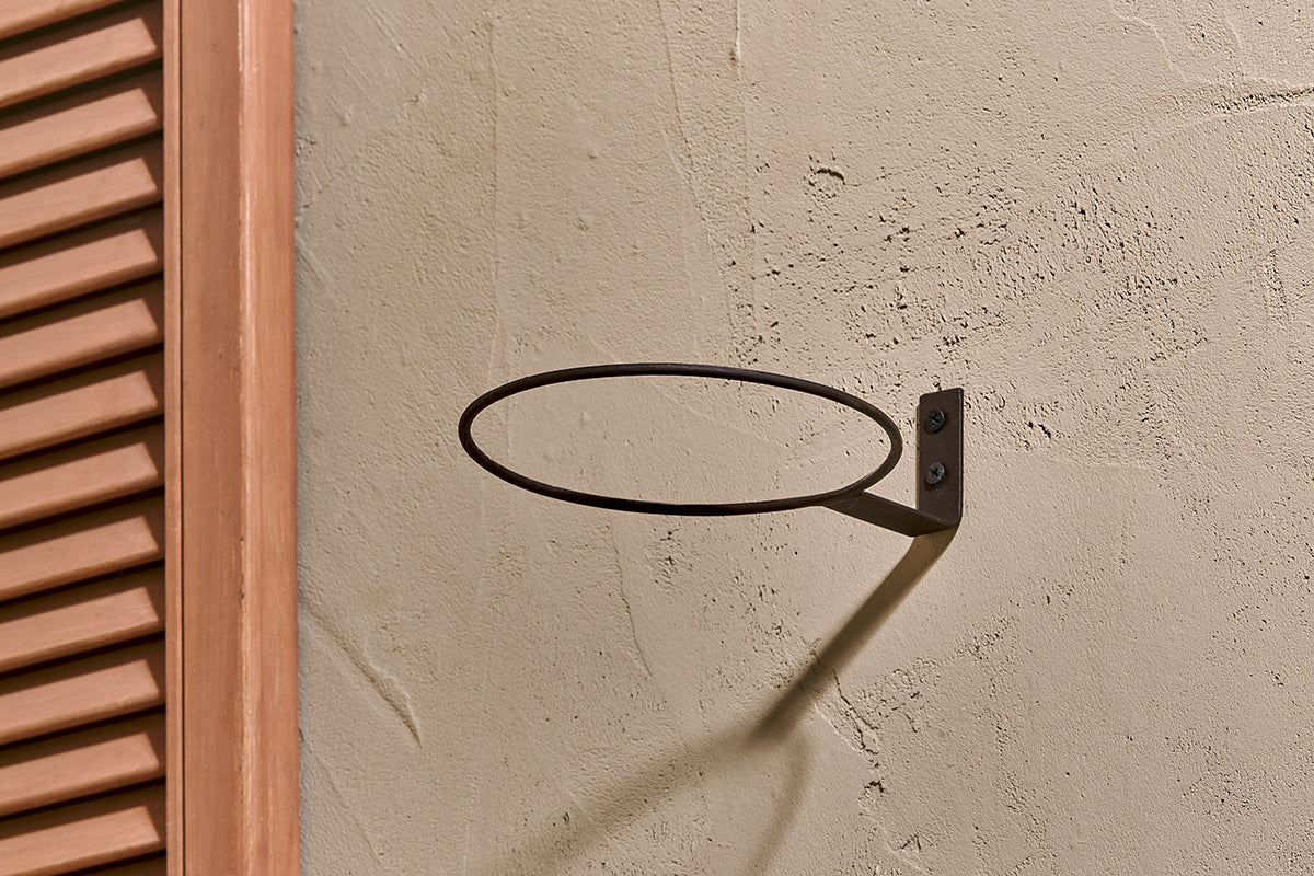 The nkuku Jara Single Wall Hung Planter, a round metal ring, is mounted horizontally on a beige textured wall with a bracket and two screws beside a wooden louvered panel. Soft natural light casts subtle shadows over the display.