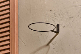 The nkuku Jara Single Wall Hung Planter is mounted on a textured beige wall, its metal ring casting an oval shadow. To the left, a partially visible brown wooden shutter with vertical slats adds contrast.