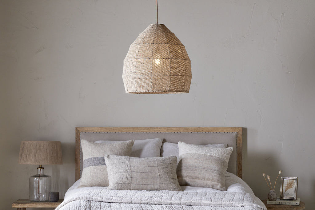 A cozy bedroom features the nkuku Jahari Woven Jute Pendant - Natural above a bed with neutral pillows and a beige blanket. Bedside tables hold a lamp, picture frame, and vase with decorative sticks.