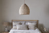 A cozy, neutral bedroom features a tufted headboard bed with textured pillows and blankets. Above, the nkuku Jahari Woven Jute Pendant in Natural adds rustic charm, while a lamp and framed art adorn the bedside table.
