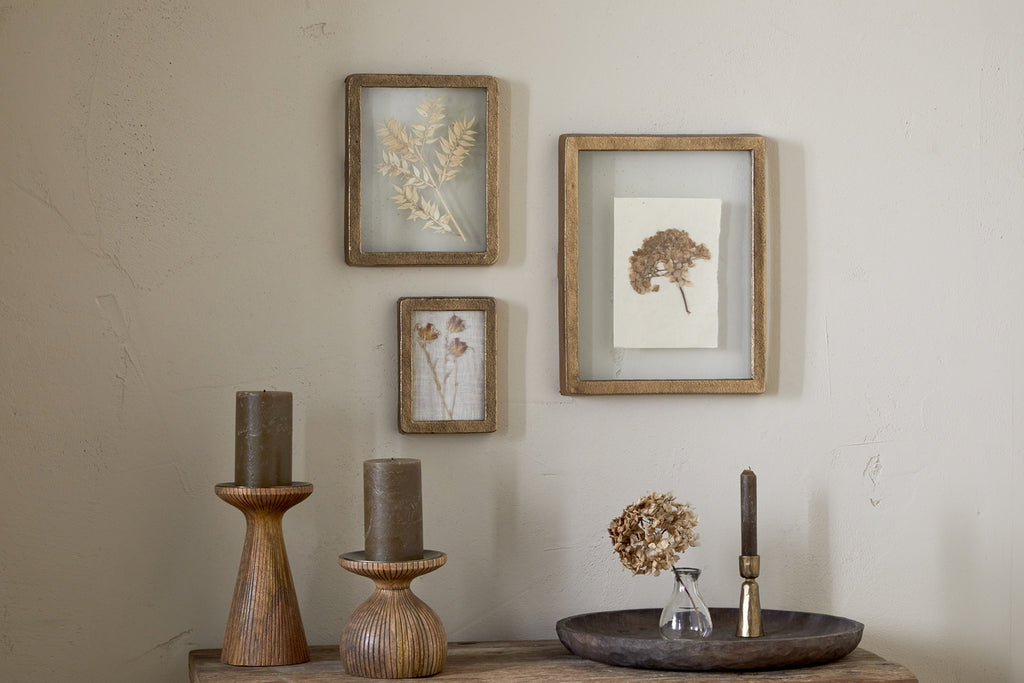 A beige wall features three nkuku Jahala Wall Hung Photo Frames in antique brass above a wooden table, which displays two brown candles, a dried flower in a glass vase, and an antique brass candle holder on a brown tray.
