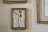 The Jahala Wall Hung Photo Frame in antique brass by nkuku hangs on a beige wall, featuring three dried flowers with faded red petals pressed against a white fabric background.