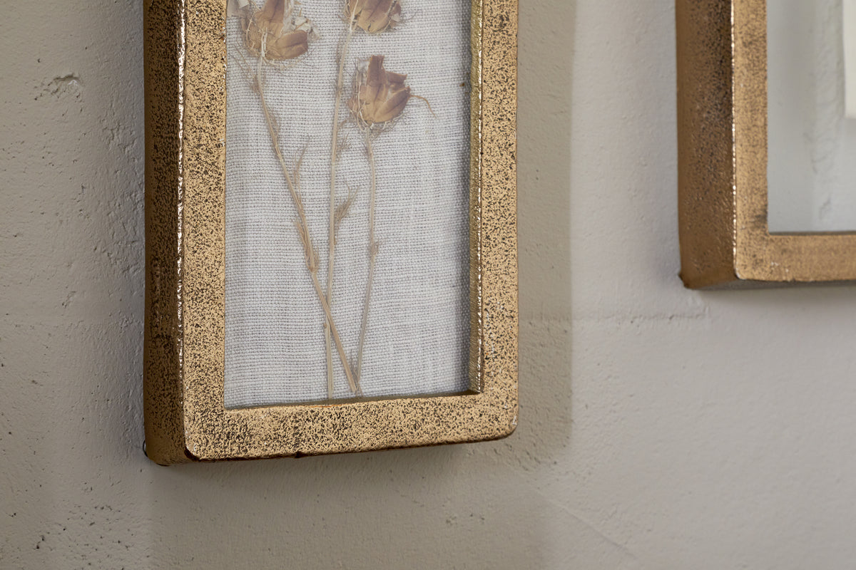 A close-up of nkuku’s Jahala Wall Hung Photo Frame in antique brass, showcasing pressed dried flowers on white mesh, hangs on a light wall, with another thick-framed photo frame partially visible to the right.