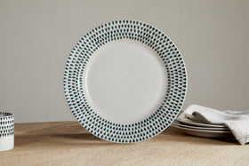 The nkuku Juniper Drop Dinnerware Set - Cream & Juniper (Set of 12) features white ceramic plates with dark blue teardrop patterns on the rim, shown here on a wooden table alongside stacked dishes, a folded beige cloth, and a matching cup.