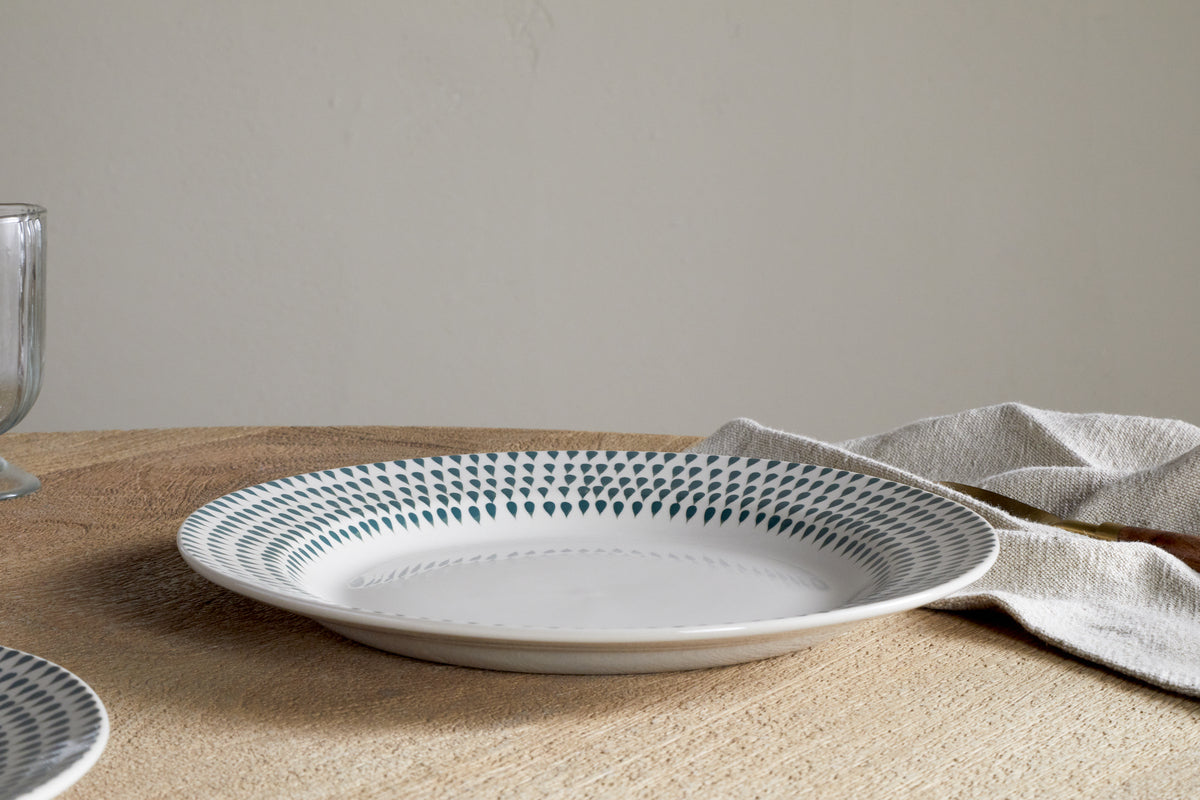 The nkuku Juniper Drop Dinner Plate in Cream & Juniper, featuring a green drop pattern on white ceramic, rests on a wooden table beside a beige linen napkin; the edge of a glass appears on the left.