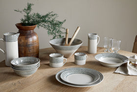 A set of nkuku Juniper Drop Dinner Plates in Cream & Juniper, featuring dark dotted patterns, is arranged on a wooden table with matching bowls, mugs, pitchers, glasses, and a brown vase with greenery in the background.