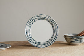 The nkuku Juniper Drop Dinnerware Set - Cream & Juniper (Set of 12) features a white ceramic plate with blue drop details on the rim, standing upright on wood, with matching bowls and a plate displayed nearby against a light wall.