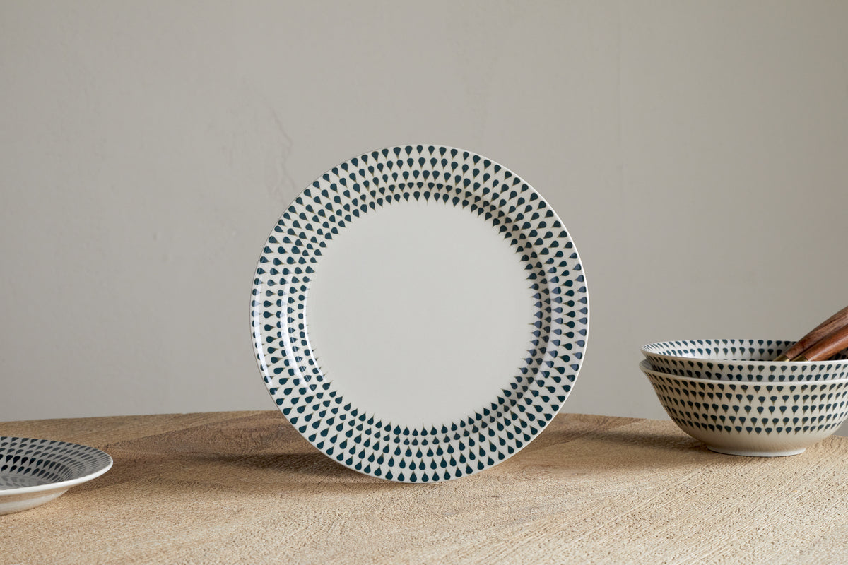 The nkuku Juniper Drop Dinnerware Set - Cream & Juniper (Set of 12) features a white ceramic plate with blue drop details on the rim, standing upright on wood, with matching bowls and a plate displayed nearby against a light wall.