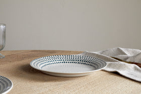 The nkuku Juniper Drop Dinnerware Set - Cream & Juniper (Set of 12) features a patterned ceramic plate with blue dots and subtle drop design on a wooden table beside a beige napkin; a glass appears partially on the left.
