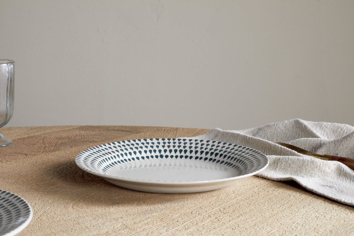 The nkuku Juniper Drop Dinnerware Set - Cream & Juniper (Set of 12) features a patterned ceramic plate with blue dots and subtle drop design on a wooden table beside a beige napkin; a glass appears partially on the left.