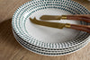 The nkuku Juniper Drop Dinnerware Set - Cream & Juniper (Set of 12) features white plates with blue-green rim accents, paired with cheese knives with wooden handles and gold blades—a stylish touch for any table.
