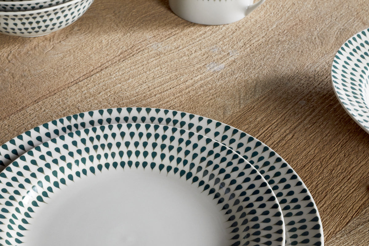 Close-up of the nkuku Juniper Drop Side Plate in Cream & Juniper, featuring a green drop pattern on the rim and set on a light wooden table, with matching dishes and a cup completing the modern dining scene.