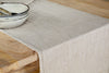 The nkuku Junto Linen Runner- Natural - Large drapes over a wooden table, while two stacked ceramic bowls with metallic silverware to the left highlight this durable table linen’s timeless elegance.