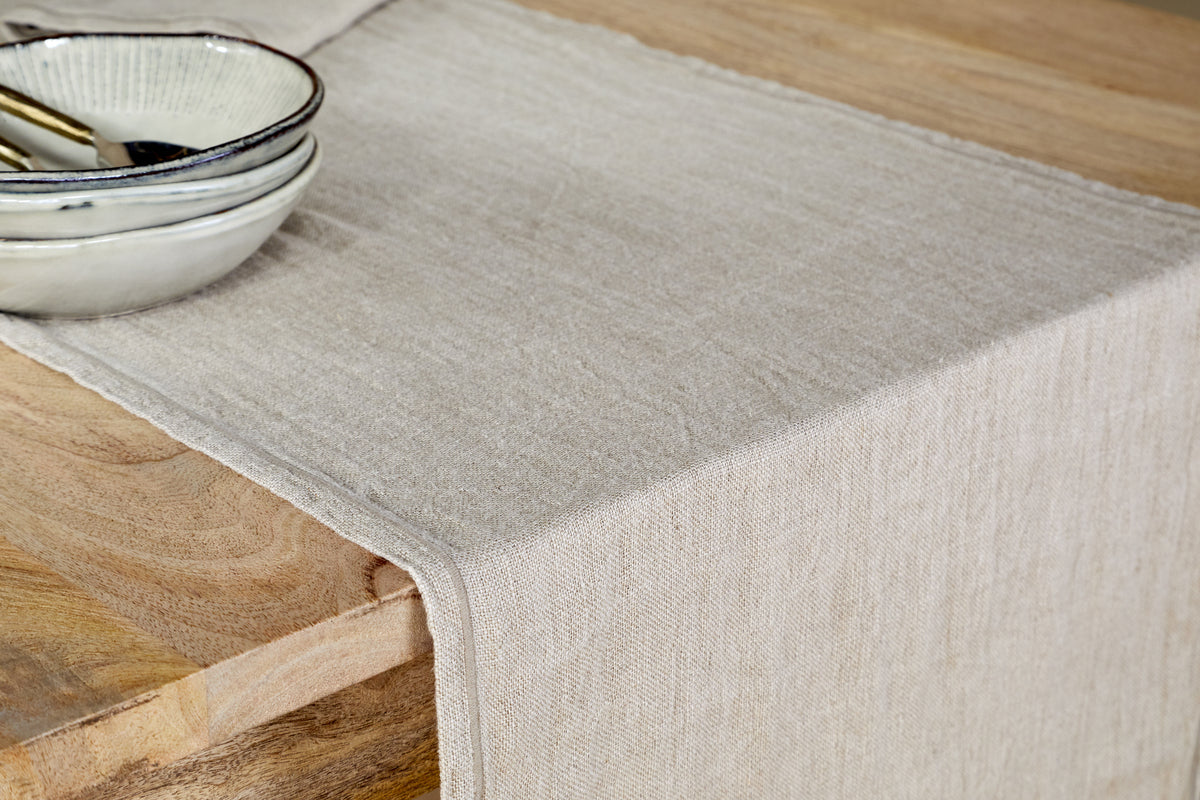 The nkuku Junto Linen Runner- Natural - Large drapes over a wooden table, while two stacked ceramic bowls with metallic silverware to the left highlight this durable table linen’s timeless elegance.