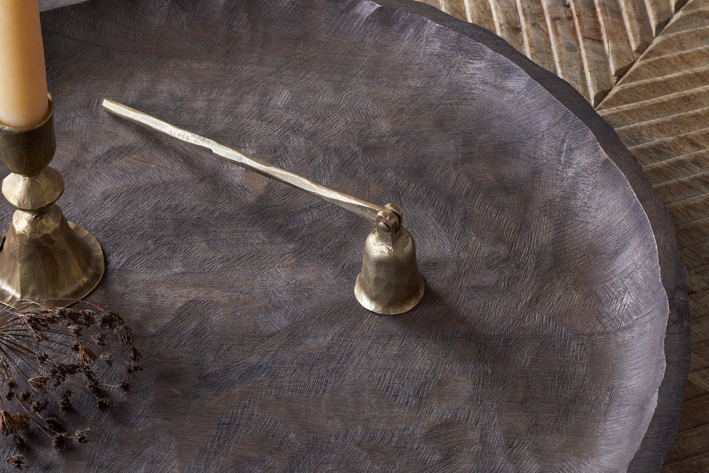 The Jyoti Candle Snuffer - Antique Brass by nkuku rests on a round, textured wooden tray with a gold candle holder and dried flowers, making it a perfect gift for candle lovers; a carved leaf pattern features in the background.