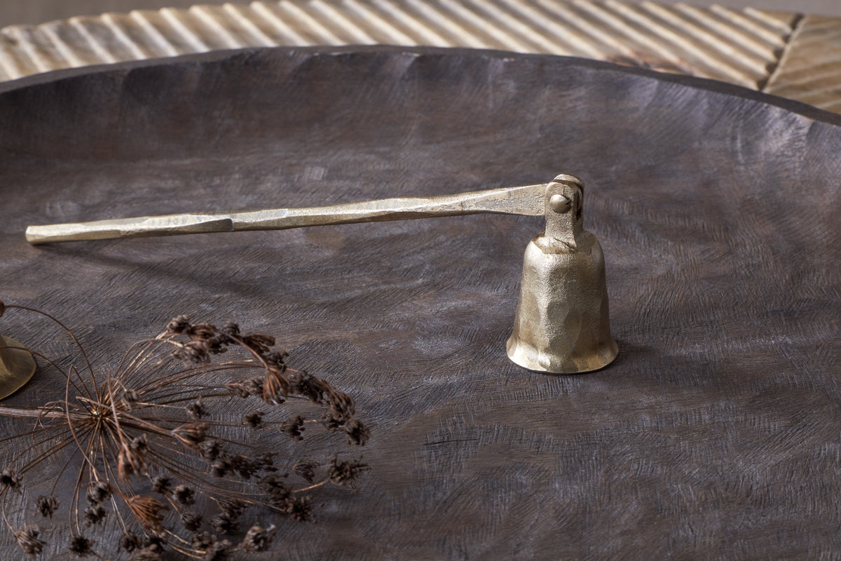 The nkuku Jyoti Candle Snuffer in antique brass, featuring a textured handle, sits on a dark metal tray beside dried botanicals—an elegant gift for candle lovers against a neutral, ridged background.