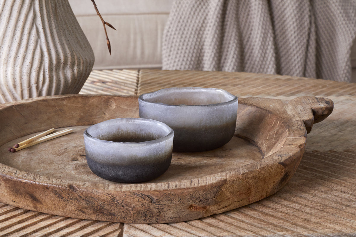 The nkuku Janka Tealight Holders - Smoked Charcoal (Set of 2) sit on a rustic wooden tray with matches, while a ceramic vase and knitted fabric in the background add to the cozy decor.