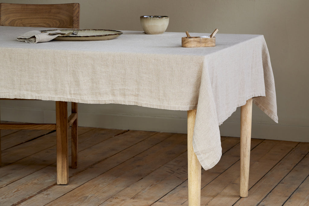 A wooden table is draped with the nkuku Junto Linen Tablecloth in Natural (Small) and set with a ceramic plate, bowl, wooden ashtray, and folded napkin. A wooden chair sits beside the table on a matching wooden floor.