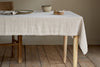 A wooden table is dressed with the nkuku Junto Linen Tablecloth in Natural (Small) and set with a ceramic bowl, small wooden container, plate, and neatly folded napkin. A wooden chair sits on the left; the floor is also wood.