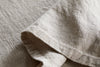 A close-up of the nkuku Junto Linen Tablecloth in Natural (Small) highlights its beige linen texture, visible weave, stitching details, and soft folds.