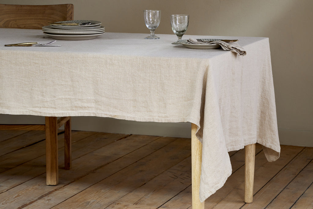 A rustic dining room with wooden floors features a nkuku Junto Linen Tablecloth in Natural (Large) draped over a wooden table, set with stacked plates, cutlery, two wine glasses, and folded napkins.