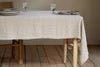 The Junto Linen Tablecloth in Natural (Large) by nkuku covers a wooden dining table set with stacked plates, glasses, and a cloth napkin. A wooden chair is partly visible on the left, and the table stands on a light wood floor.