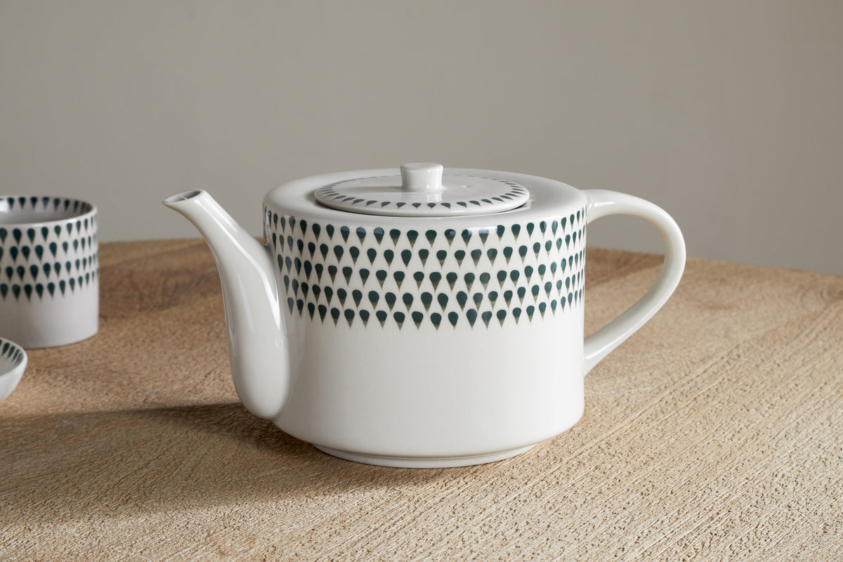 The nkuku Juniper Drop 4 Person Teapot in Cream & Juniper, featuring a round handle and spout and decorated with dark teardrop motifs, sits on a light wood surface—an elegant hand-painted gift for tea lovers, with a matching cup behind.