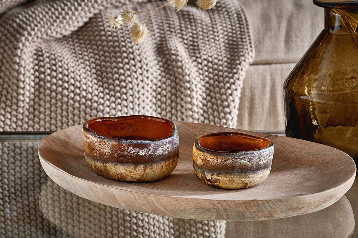 The Janka Tealight Holders - Dark Amber (Set of 2) by nkuku sit on an oval wooden tray, with a chunky beige knit blanket draped over a couch and a recycled glass vase nearby, creating a cozy, earthy setting.