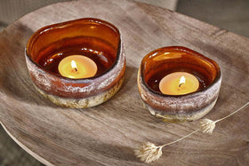 Two lit Janka Tealight Holders - Dark Amber by nkuku, crafted in rustic brown ceramic with uneven edges, sit atop a round wooden tray. A single dried grass stem nearby adds a natural accent to the cozy display.