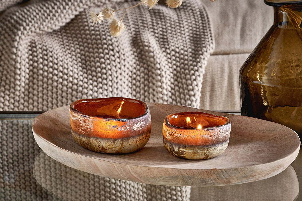Two nkuku Janka Tealight Holders in dark amber, set of 2, glow atop a wooden tray on a glass table. A beige knitted blanket and brown recycled glass vase add to the space's warm, rustic charm.