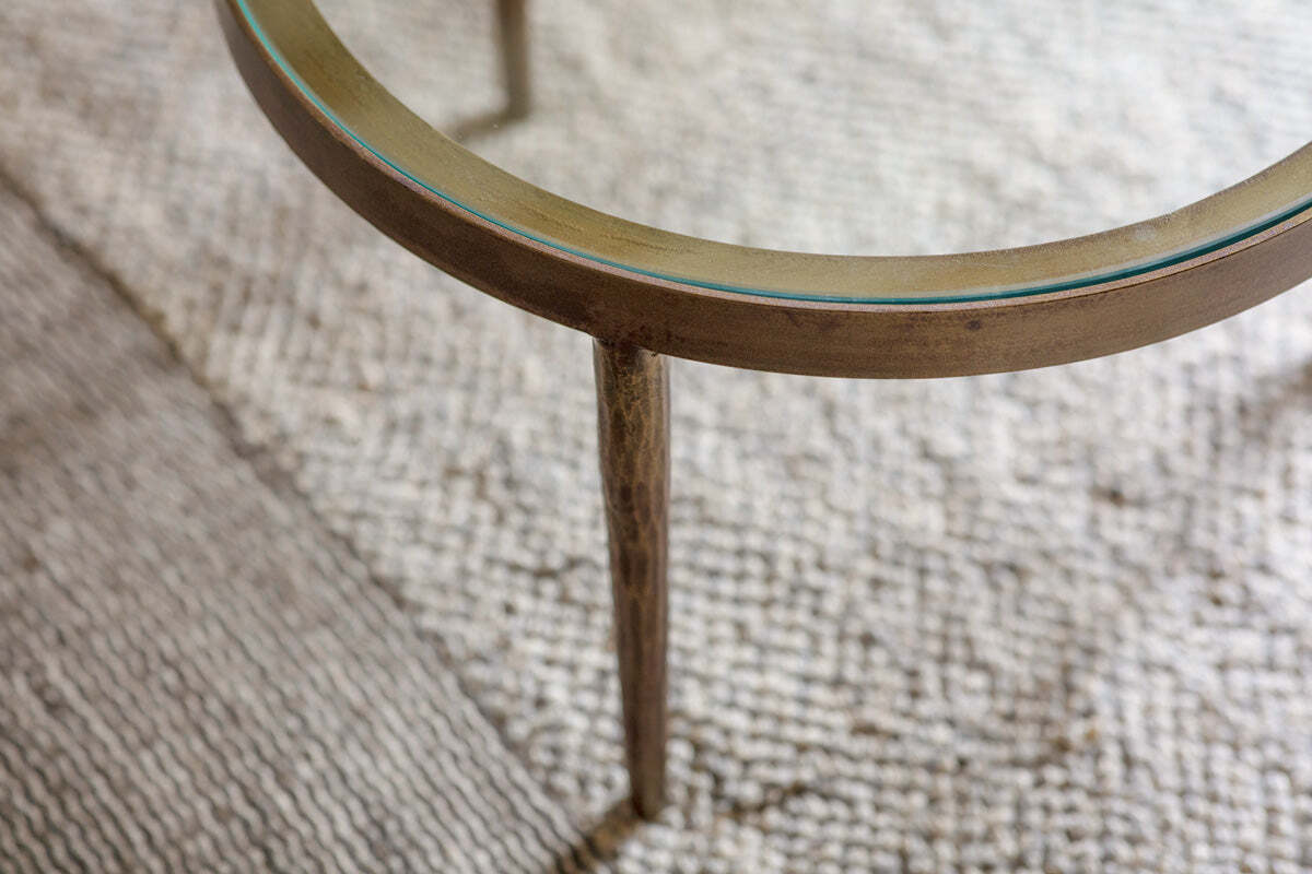 Close-up of an nkuku Jiya Iron & Glass Nested Coffee Table’s round, slender brass-finished leg on a textured beige rug, highlighting the metallic sheen—ideal for pairing with glass tops.