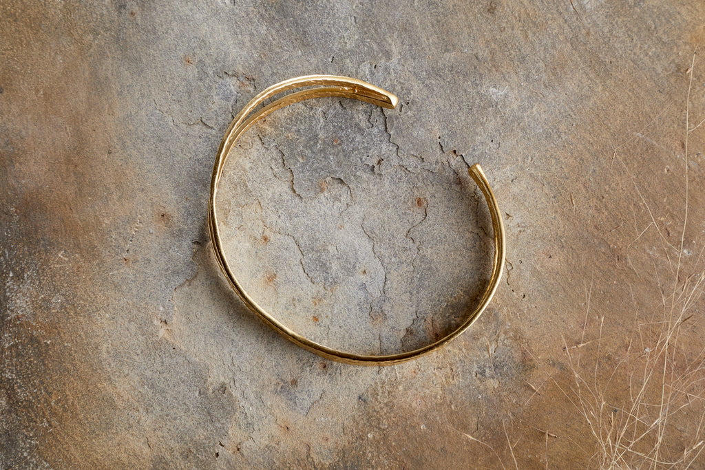 The Katai Bangle - Gold by nkuku, featuring open ends and a subtle hammered texture, rests on a textured, cracked stone surface.