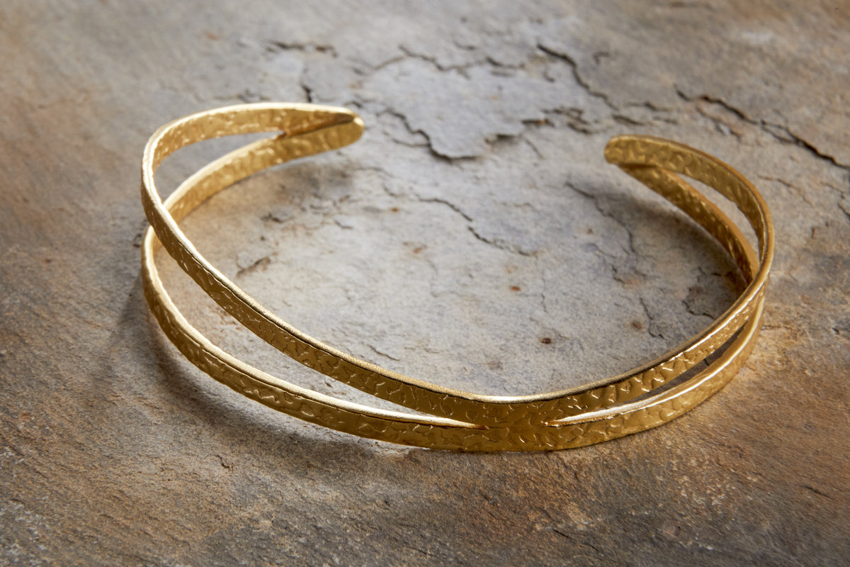 The Katai Bangle - Gold by nkuku features a sculptural, hammered double band design that gleams against a textured, weathered stone surface.