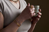 Wearing a beige top and sculptural jewelry, a person holds a ceramic mug with both hands. On their wrist gleams the Katai Bangle - Gold by nkuku. The background is dark and plain.