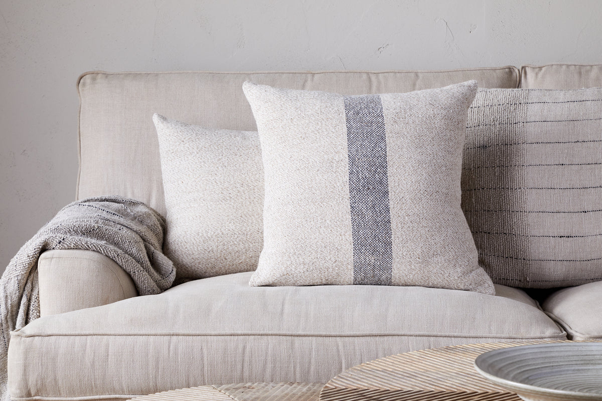 A beige sofa features the nkuku Kallu Wool & Cotton Cushion Cover in natural, a textured cream cushion, and a gray throw blanket. A light background wall and woven tray on the table finish this serene setting.